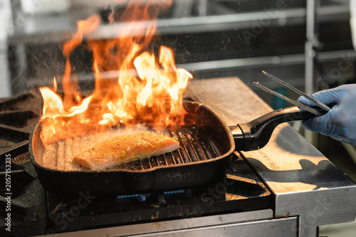 Seared salmon fish cooks in flaming cast-iron pan. Flames leap from grilling pan. Salmon fish cooks intensely. High-heat grilling action. Steam rises from sizzling fish.