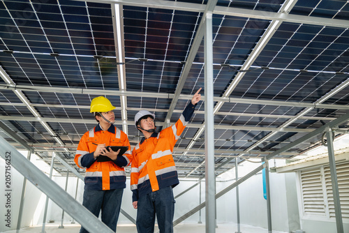Young technician and Engineer inspect solar panels on steel framework on building rooftop. One points upward, the other holds a tablet. They wear bright orange safety vests and hard hats.