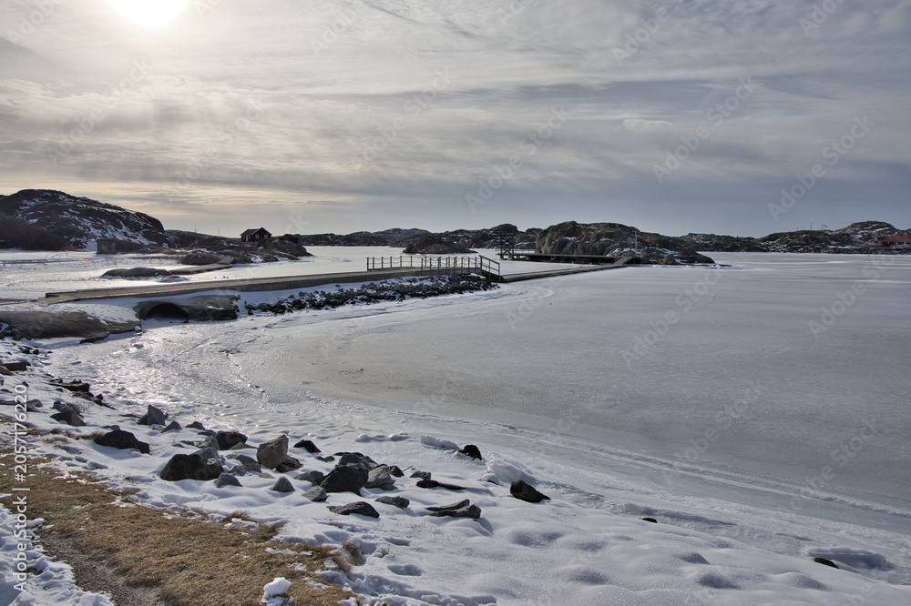 Naklejka premium Symmetrical view of a wooden pier with rope railings leading across the ice in Skrhamn. A cold winter day in the Swedish archipelago with dramatic light and frozen wate