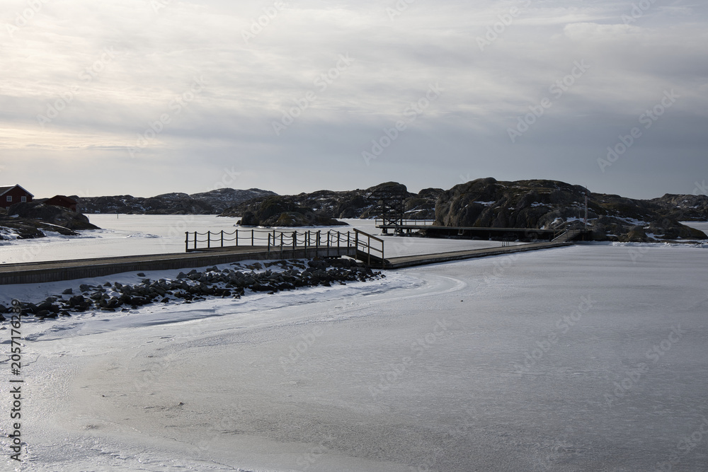 Naklejka premium Symmetrical view of a wooden pier with rope railings leading across the ice in Skrhamn. A cold winter day in the Swedish archipelago with dramatic light and frozen wate