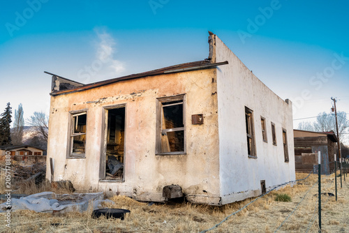 An old stucco building stands against a blue sky. The exterior walls are discolored and the windows and doors are long gone.