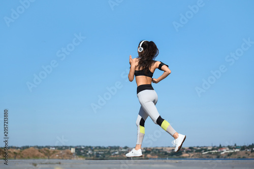 Young sporty Asian woman in headphones jogging along waterfront promenade out...