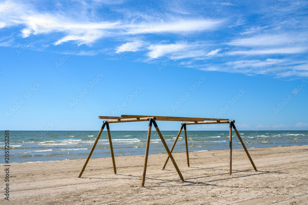 Fototapeta premium Empty wooden canopy frame setup on a sandy beach next to the sea under a bright blue sky with wispy clouds in summer.
