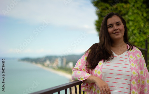 Travel city photo. Female portrait. Young adult white woman with pink kimono dress, t-shirt with stripes in spring summer park looks at camera. View of sea beach, blue sky. Concept of nature, rest