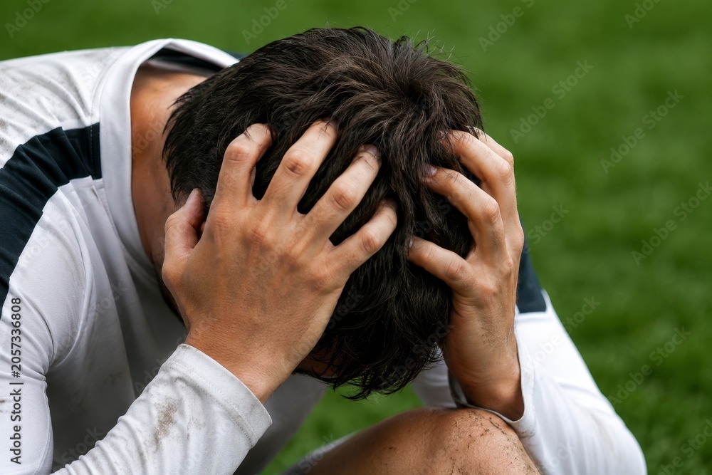 Fototapeta premium Soccer player holding head in hands on grass field after losing match. Upset athlete wearing white uniform during game. Sports defeat, failure, disappointment and sadness concept.