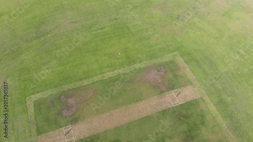On command drone camera descending, tilting over grassy field revealing cricket pitch and stumps