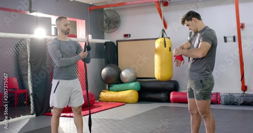 After demo, white coach and indian trainee wrapping black, red hand wraps on mat for sparring