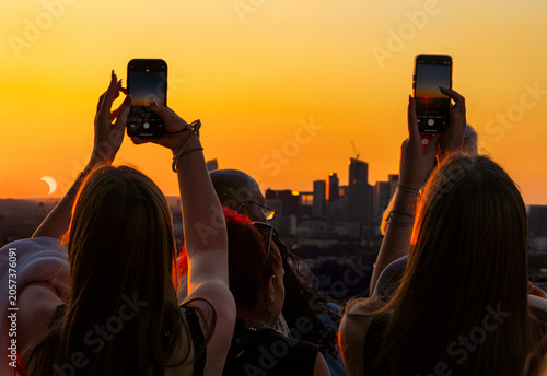 Backlit people, from the top of the Eiffel Tower, capturing a beautiful partial solar eclipse with their smartphones against a golden sunset sky with the Paris skyline and La Défense in the background