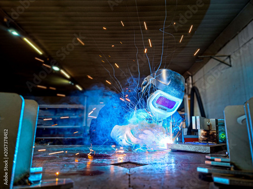 A wide-angle industrial shot of a welder working at a metal table. Brilliant blue arc light and smoke surround the worker.