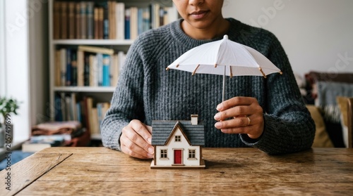 Person holding umbrella over miniature house symbolizing home insurance protection.