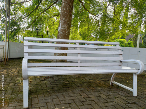 White painted metal bench with wooden slat design placed under a large tree shade on paving block floor. Outdoor park or garden furniture background detail.