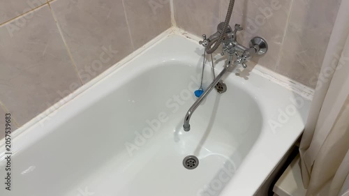 White bathtub with chrome faucet close-up, economy hotel bathroom empty tub with handheld spout dripping, tiled walls and shower
