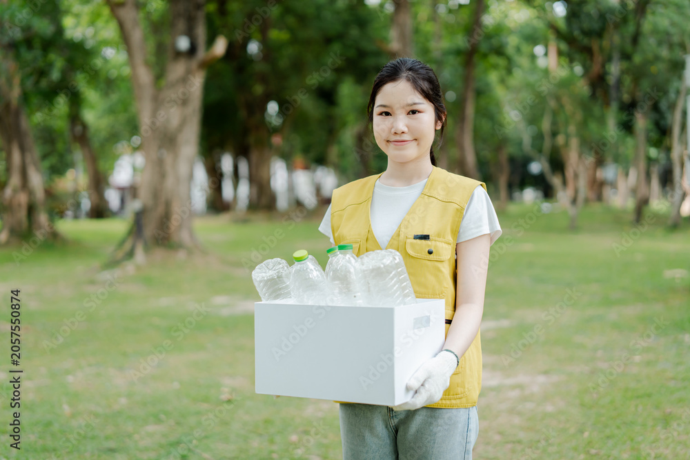Fototapeta premium Young asian woman volunteer holding a box with plastic bottles, participating in a recycling program and contributing to environmental sustainability in a park