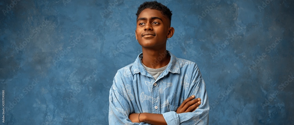 Naklejka premium A confident young man stands with crossed arms against a textured blue background, embodying modern youth culture and self-assurance.