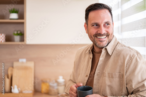 Smiling man finding joy and relaxation in his bright kitchen, holding a mug of coffee and looking out, embodying a serene and happy start to the day at home