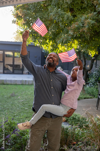 Mature African American father holding child and waving American flags in yard wearing blue shirt