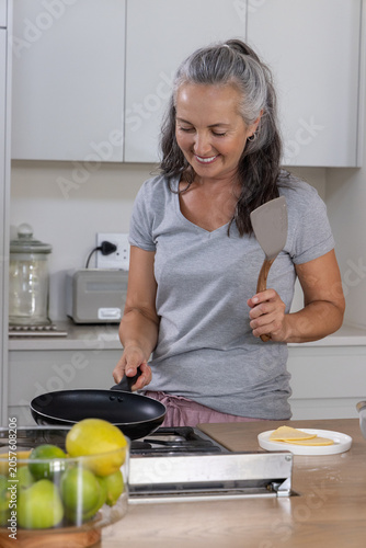 Middle-aged woman standing at stove preparing food with nonstick pan and spatula in kitchen