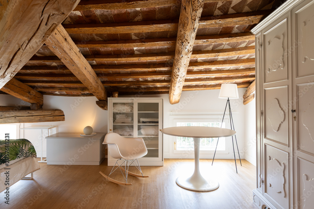 Obraz premium In the bedroom with exposed beams, there's this space with a round table and a lamp. Behind it, a dresser with blankets and a light bulb.