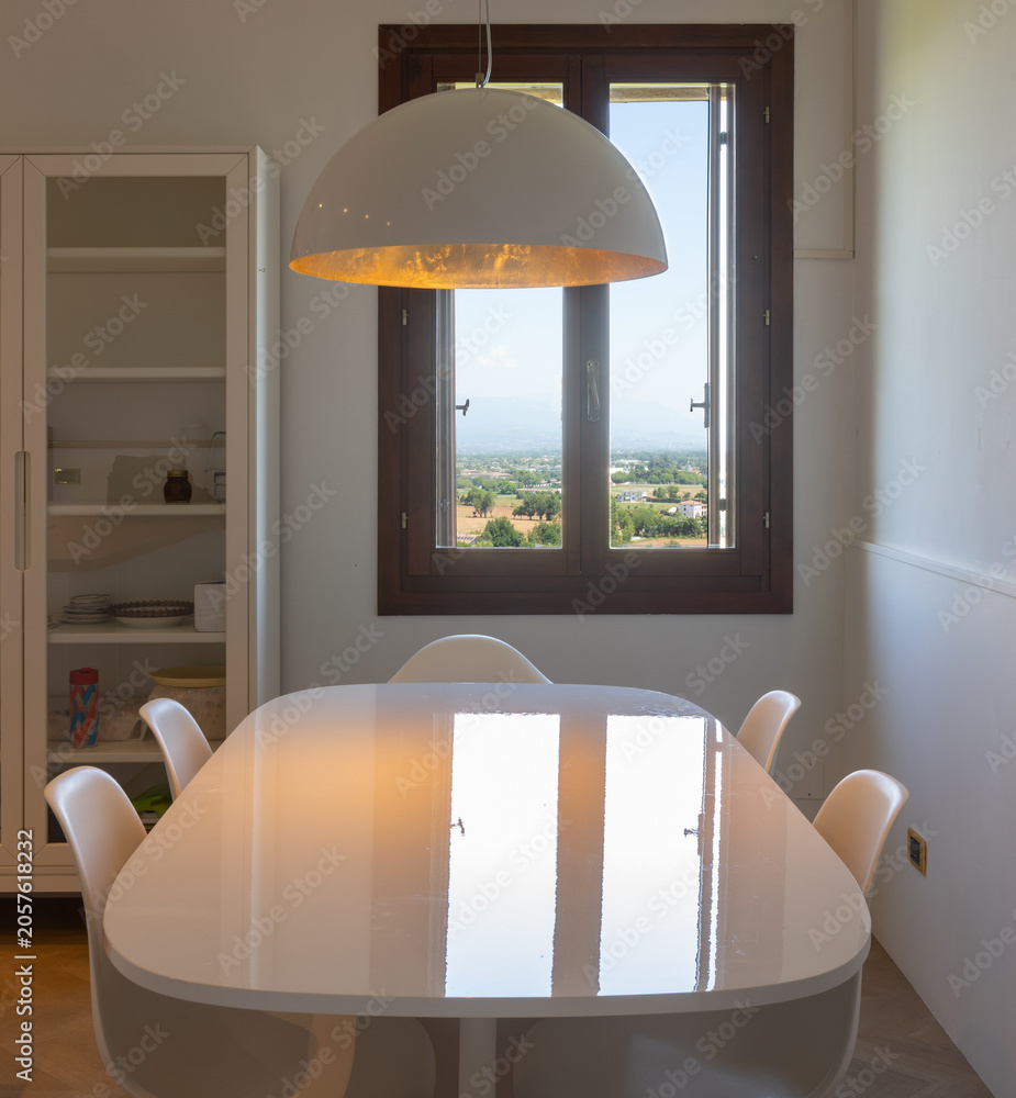 Obraz premium Front view of an oval white table, with chairs underneath, a sideboard to the left, and a window overlooking the Italian hills. Country house