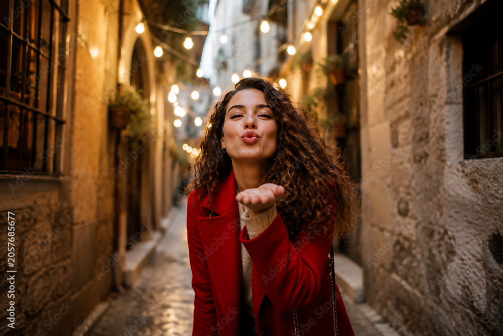 Obraz premium Young Woman Blowing Kiss In Charming Historic European Street With Warm Lights Romantic Travel Lifestyle Portrait Photography Concept