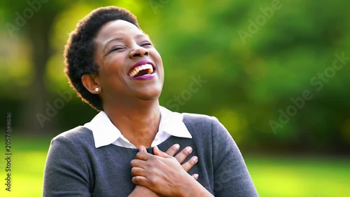 African american woman, mid-30s, business-casual top, laughs and triumphs, then smiles with gratitude. warm golden hour light, lush green outdoor bokeh, medium video, concept of success and profound