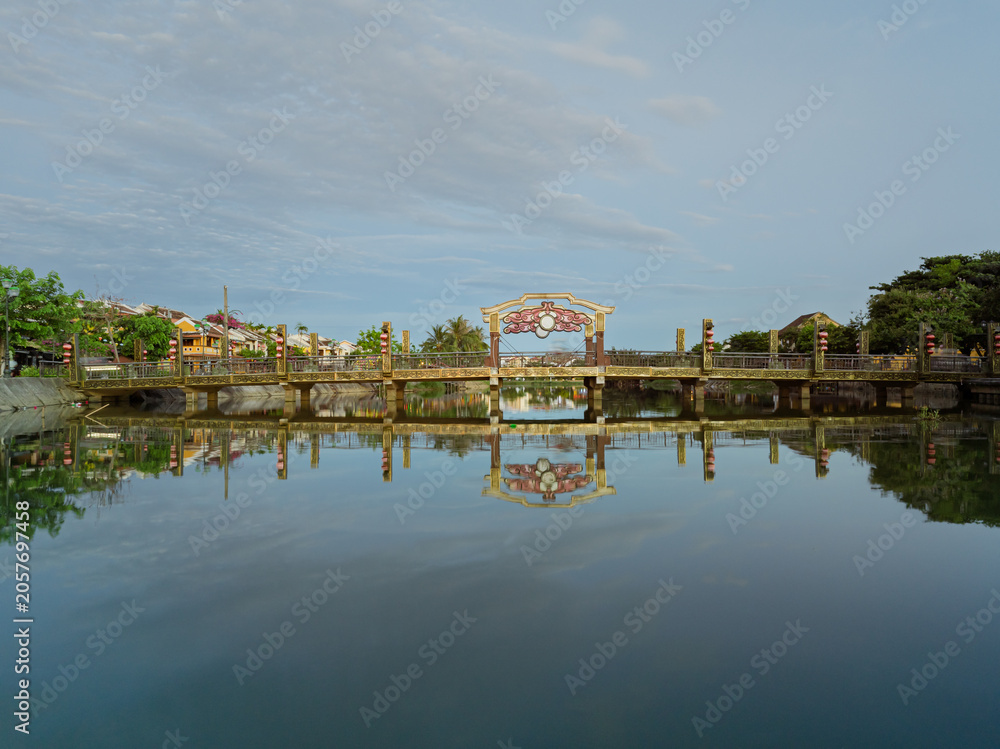 Fototapeta premium Historic bridge reflected in calm water along a peaceful canal in Hoi An Ancient Town.