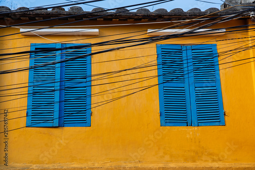 Blue wooden shutters contrast with a yellow heritage wall in Hoi An Ancient Town, Da Nang, Vietnam.
