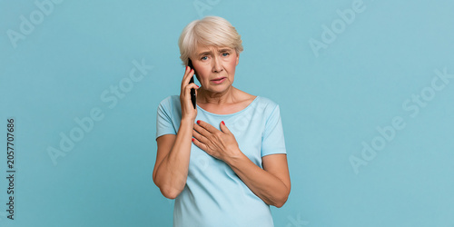 An elderly woman with short, white hair talking on a mobile phone with a concerned expression. She is wearing a light blue t-shirt and has her hand on her chest, suggesting distress or discomfort