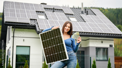 Woman holding solar panel and showing money near house with rooftop panels. Concept of renewable energy savings, eco friendly lifestyle, sustainable home