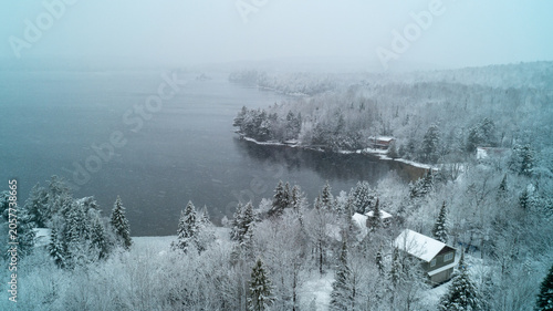 A moody high-angle drone photograph over a calm northern lake shoreline during a heavy winter snowfall, showcasing snow-covered evergreen trees and waterfront cottages under a hazy, overcast sky.
