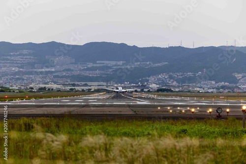 Fotografie 飛行機 滑走路 空港の風景。夕暮れ時の滑走路から離陸または着陸する航空機の後ろ姿と、点灯する誘導灯、背景に広がる山々と街並み、旅や輸送をイメージする景観素材。