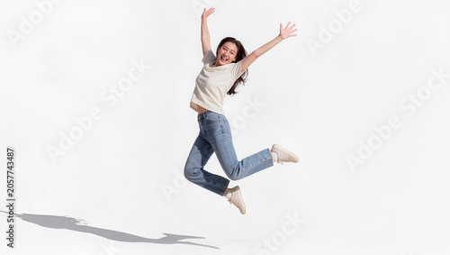 Joyful young Asian woman jumping in mid air with arms raised on white background, freedom and happiness lifestyle concept, editorial portrait photography with copy space