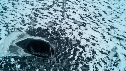 Quadro su tela A striking top-down drone photograph looking straight down at a partially frozen winter lake, featuring a dramatic deep black open ice hole surrounded by complex white snow and slush patterns