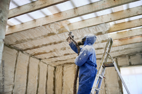 Professional worker applies expanding foam insulation to timber structure from ladder. Unfinished home interior, protective mask, building performance and home improvement concept
