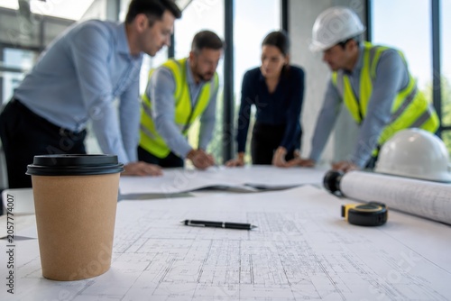 Group of construction professionals collaborating on blueprints at a table with a coffee cup in the foreground, wearing safety vests and hard hats in a modern office setting