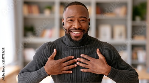 Happy Black man smiles with hands over his heart. He wears a dark ribbed turtleneck in a cozy room with shelves behind him. His joyful expression suggests sincerity or gratitude