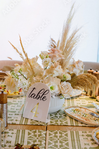 A close-up of an elegant wedding banquet table centerpiece featuring a boho floral arrangement in a white vase and a golden Table 4 sign.
