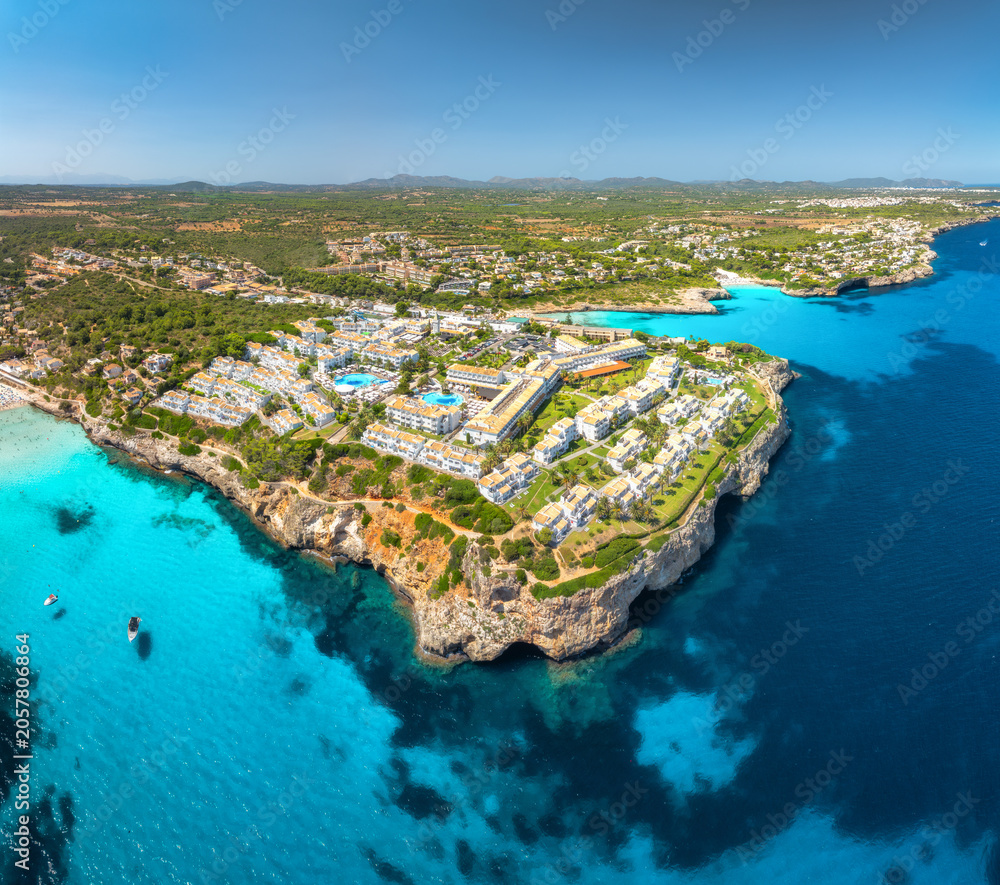 Obraz premium Aerial drone panorama view of sea coast with white resort houses on rocky cliffs. Turquoise blue sea water, sunny sky, boats, sandy beach. Luxury travel destination, Mallorca island, Spain. Top view