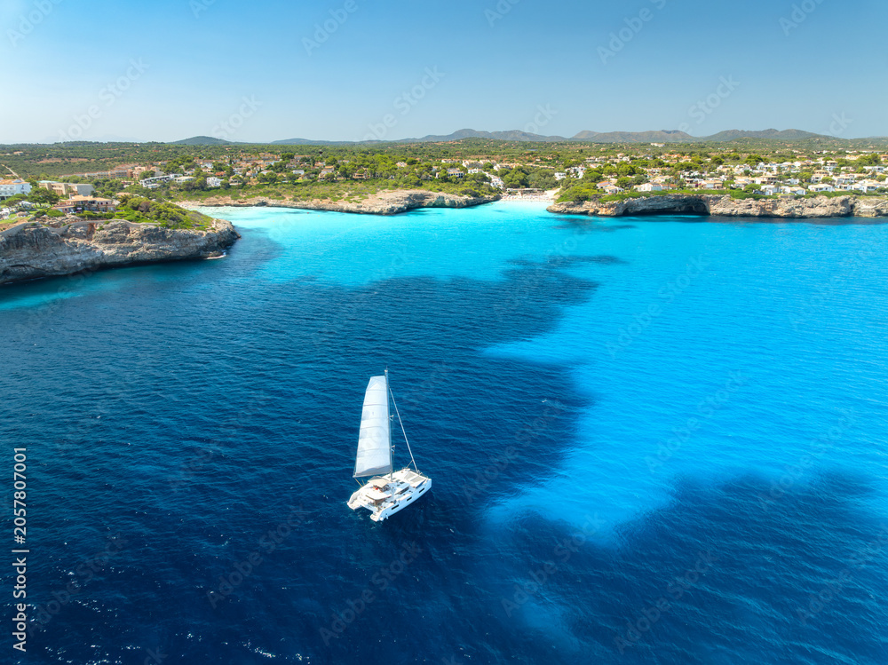 Obraz premium Aerial view of sailing catamaran yacht in turquoise sea water bay. Sailboat. Mallorca, Spain. Luxury boat cruising in ocean cove near sandy beach, resort houses on rocky coast. Drone summer landscape