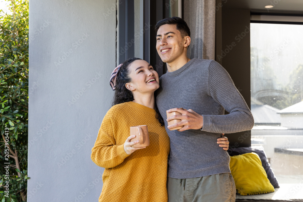 Obraz premium Couple standing in doorway leaning together holding ceramic mugs by sofa and sliding glass door