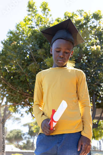 African American child wearing black cap yellow top holding red-ribbon diploma looking down in park