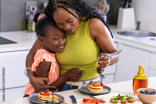 African American mother in green hugging youth daughter in peach at countertop with pancakes, fork