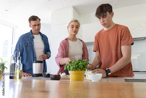 Family with parents and teenage son preparing food at kitchen island using lemon, blender, basil