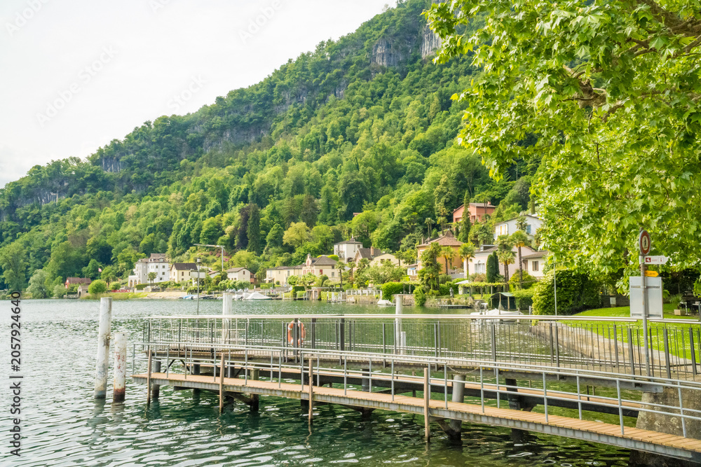 Fototapeta premium Wooden pier on Lake Lugano with village and forested mountain Caslano Switzerland