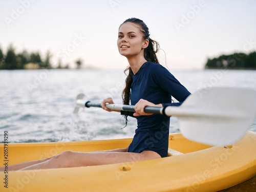 Фотография An athletic woman sits in a yellow kayak on calm water, paddling with a double blade paddle, smiling toward the camera and enjoying a sunny outdoor moment