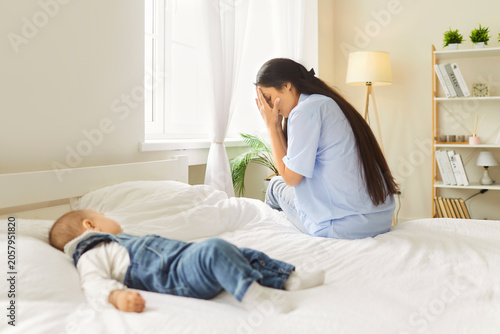 Tableau sur toile A tired mother sits on the bed covering her face while her baby lies nearby on white sheets in a bedroom, showing exhaustion and postpartum strain