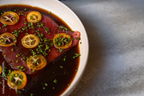 A plate holds a fresh tuna dish prepared with a dark sauce. Slices of citrus fruit and herbs are placed on top. This is happening in a restaurant setting, showcasing the food presentation.