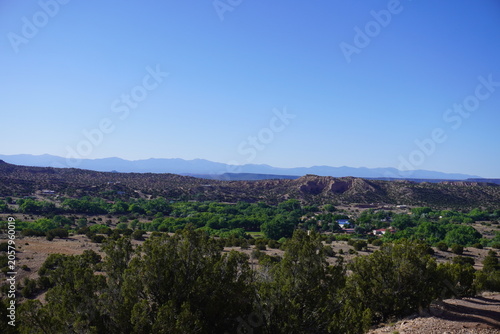 A wide angle panoramic shot of a lush green river valley oasis cutting through a rugged desert landscape, with dense trees and scattered houses nestled below rolling arid hills and a distant mountain 