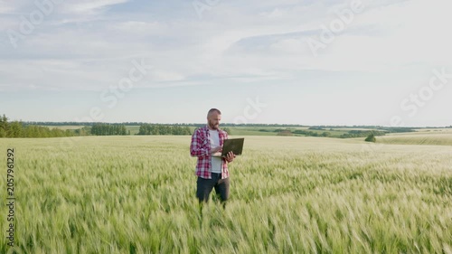 A young male farmer stands in the middle of a vast green wheat field under a blue sky.The agronomist is holding an open laptop to analyze and monitor the crop growth.