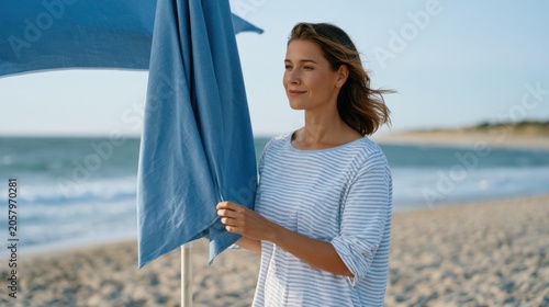 Young woman in striped shirt adjusts blue beach umbrella on sandy shore, ocean waves and clear sky visible in the background during peak summer vacation